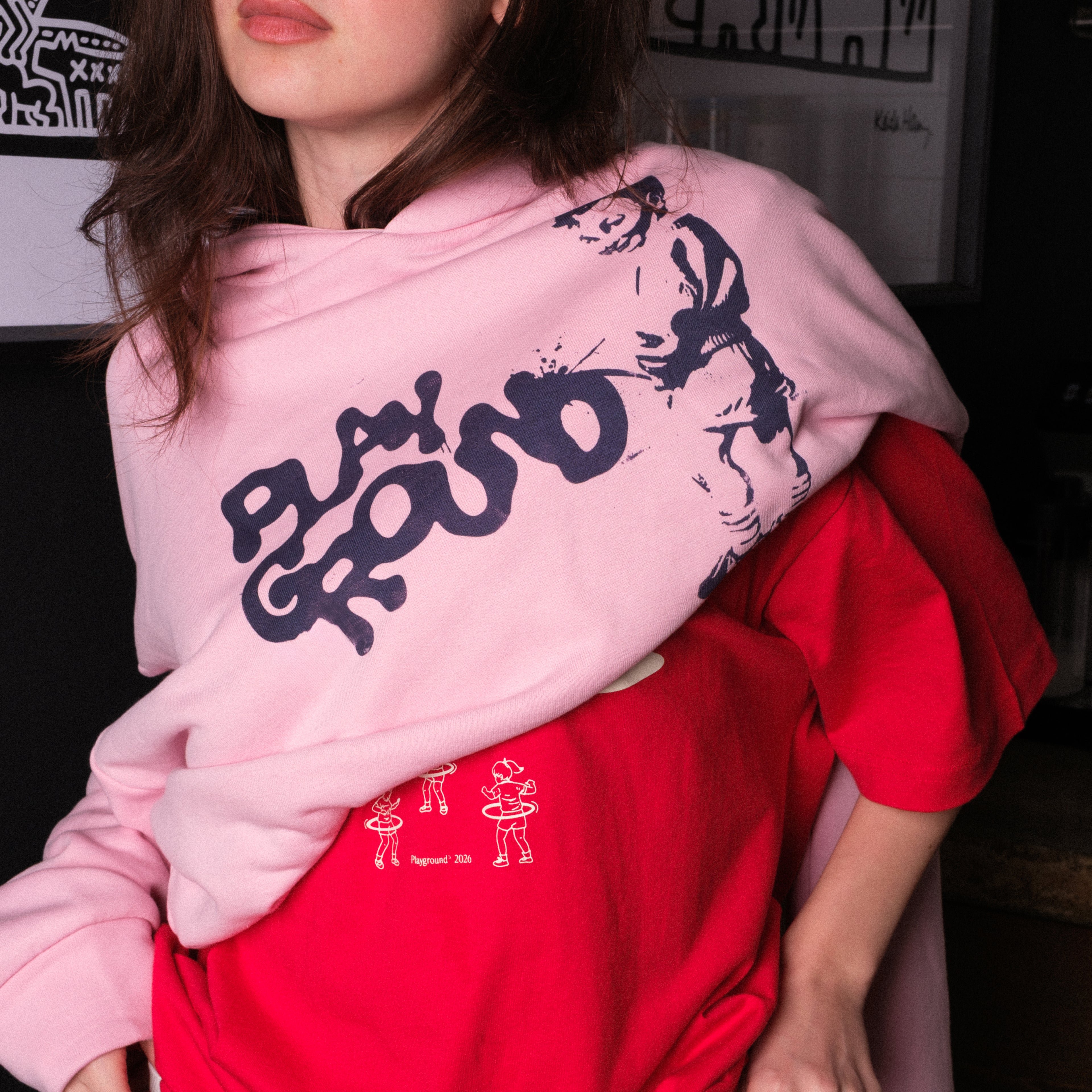 Model wearing a pink hoodie with black text and graphics, sitting against a dark background.