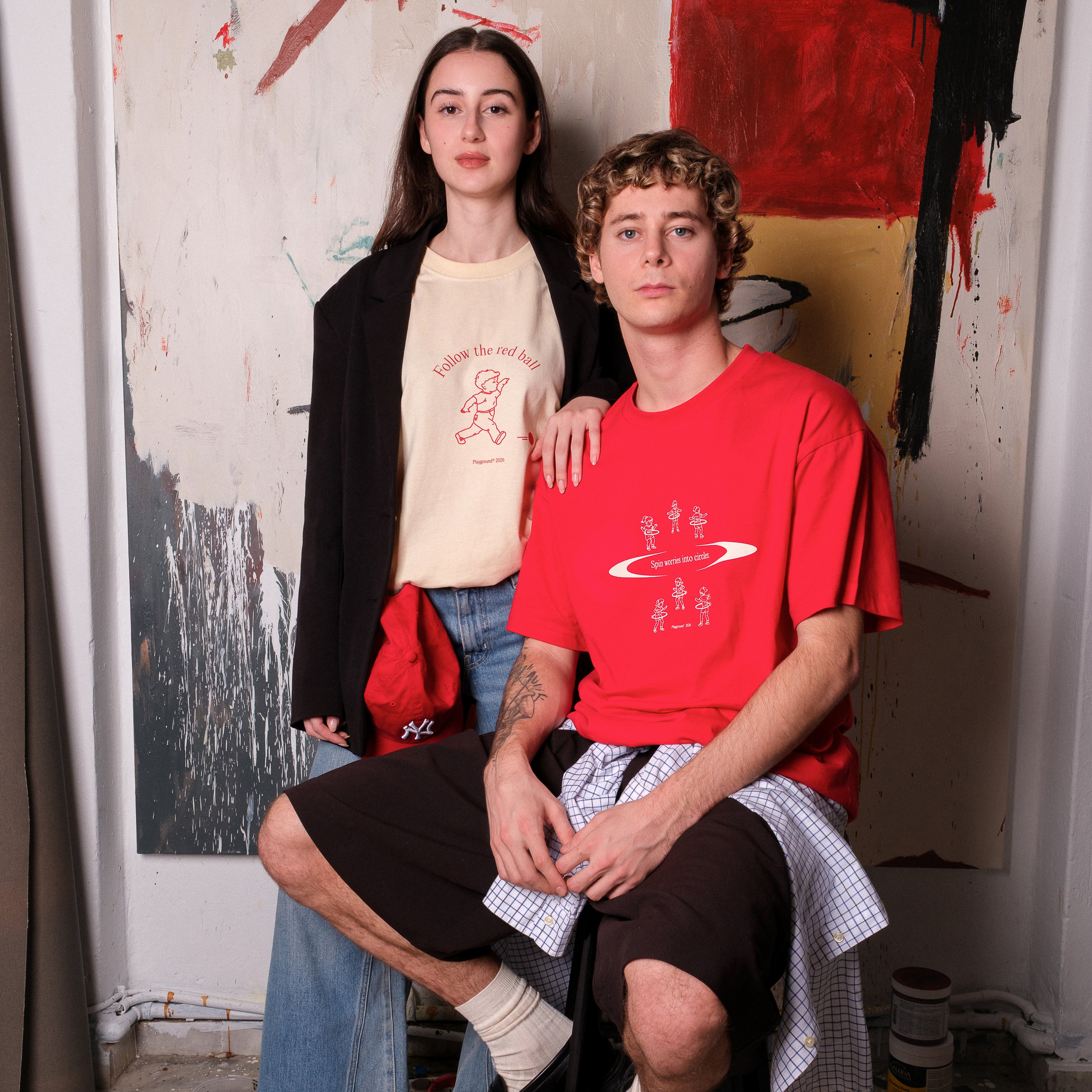 Two models people wearing t-shirts with graphic designs in a room with artistic wall decorations. One tshirt says "SPIN WORRIES INTO CIRCLES" while the other says "FOLLOW THE REDD BALL"